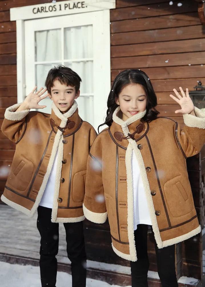 Family of four wearing matching brown shearling coats with white accents, posing outside a rustic log cabin in snow